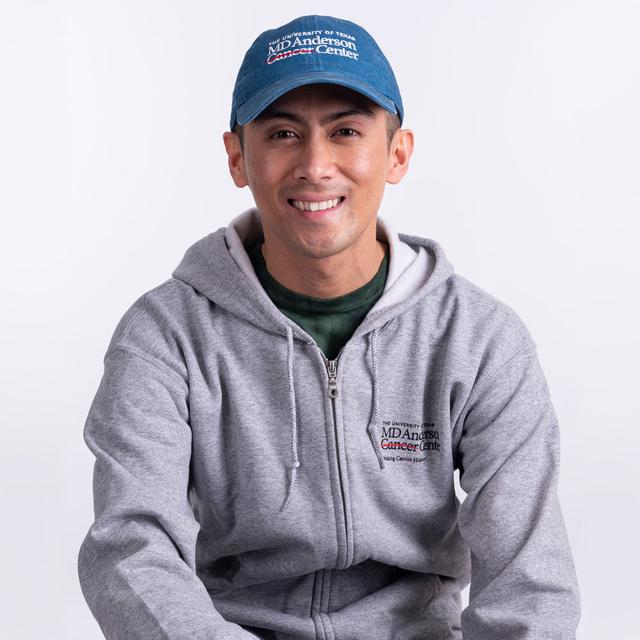 MD Anderson employee wearing a grey zippered hoodie with pockets, featuring the black MD Anderson logo on the chest area, paired with a denim baseball cap with the white MD Anderson logo.